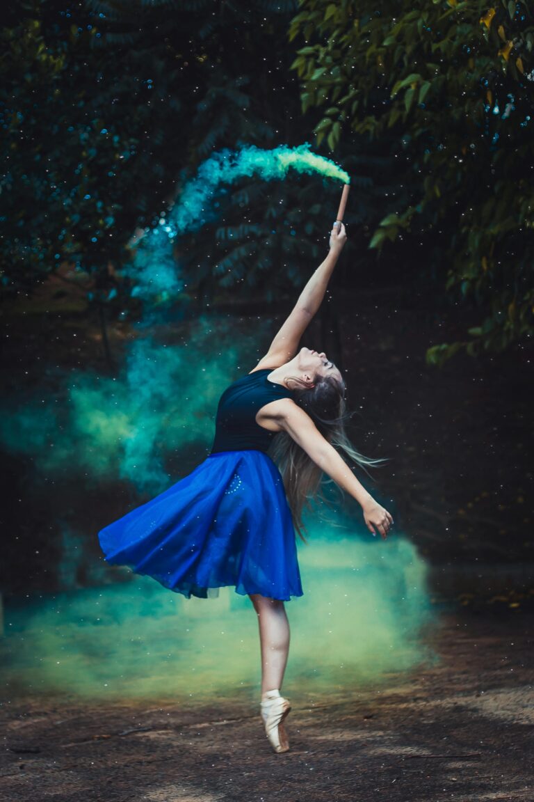 A graceful ballerina in a blue dress performs an elegant dance with colored smoke, showcasing artistic flair.